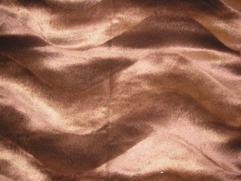 Falista, brązowa tkanina z delikatnym połyskiem, przypominająca aksamit lub welur. Widoczne subtelne załamania i cienie podkreślające fakturę materiału.