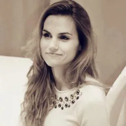 Portret uśmiechniętej kobiety z długimi włosami, ubrana w jasną bluzkę z ozdobnymi kamieniami przy dekolcie, spoglądającej w lewo.