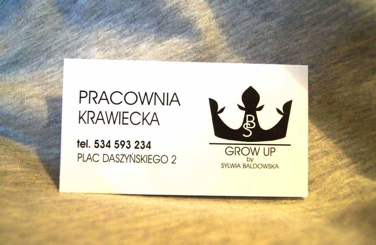 Wizytówka pracowni krawieckiej z czarnym logo korony i napisem 'GROW UP by Sylwia Baldowska', leżąca na szarym tle. Zawiera numer telefonu i adres.