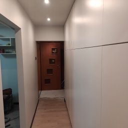 J-Rem Jacek Sławiński - Korytarz po remoncie: białe ściany, zabudowa szafy wnękowej, drewnopodobne panele podłogowe, brązowe drzwi z dekoracyjnymi aplikacjami. Widoczne narzędzia.