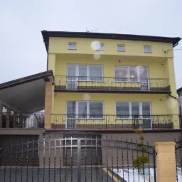 Żółty, dwupiętrowy dom z balkonami i zadaszonym miejscem parkingowym, widziany zza czarnego, kutego ogrodzenia. Widoczne detale architektoniczne i sąsiednie budynki.