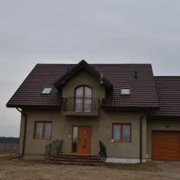 Dwukondygnacyjny dom z brązowym dachem i garażem, widok z przodu, schody wejściowe z balustradami. Szara elewacja, brązowe okna i drzwi. Pochmurne niebo.