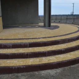 Żółto-brązowa kostka brukowa ułożona w łukowate schody i okrągły wzór na podjeździe, widok z dołu, na tle szarej elewacji budynku i ogrodzenia.