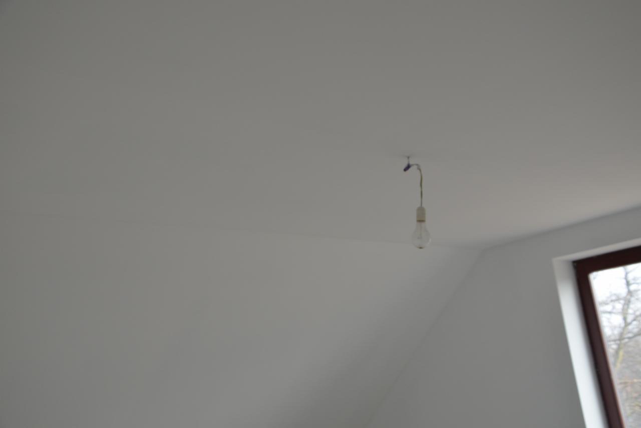 Biały pokój na poddaszu z oknem i żarówką wiszącą na kablu. Minimalistyczne wnętrze w trakcie wykańczania. Widoczny skos dachu i fragment drzewa za oknem.