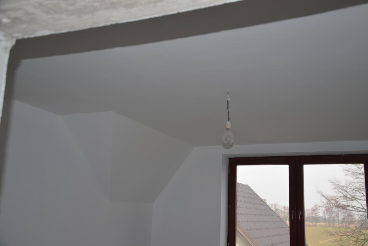 Wnętrze w trakcie malowania: białe ściany, sufit z żarówką na kablu, okno z widokiem na zewnątrz. Widoczny fragment betonowej konstrukcji sufitu.