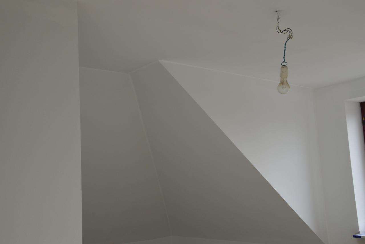 Białe, świeżo pomalowane poddasze ze skosem, z wiszącą żarówką na kablu. Widoczne okno i kąt ściany. Minimalistyczne wnętrze po remoncie.