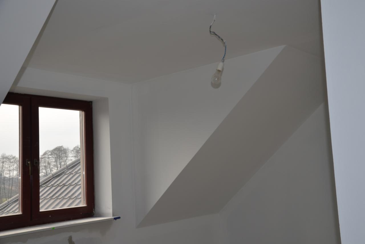 Poddasze po malowaniu na biało, widoczne okno z brązową ramą i surowa żarówka na kablu. Ściany gładkie, bez dekoracji. Widok na dach budynku i drzewa za oknem.