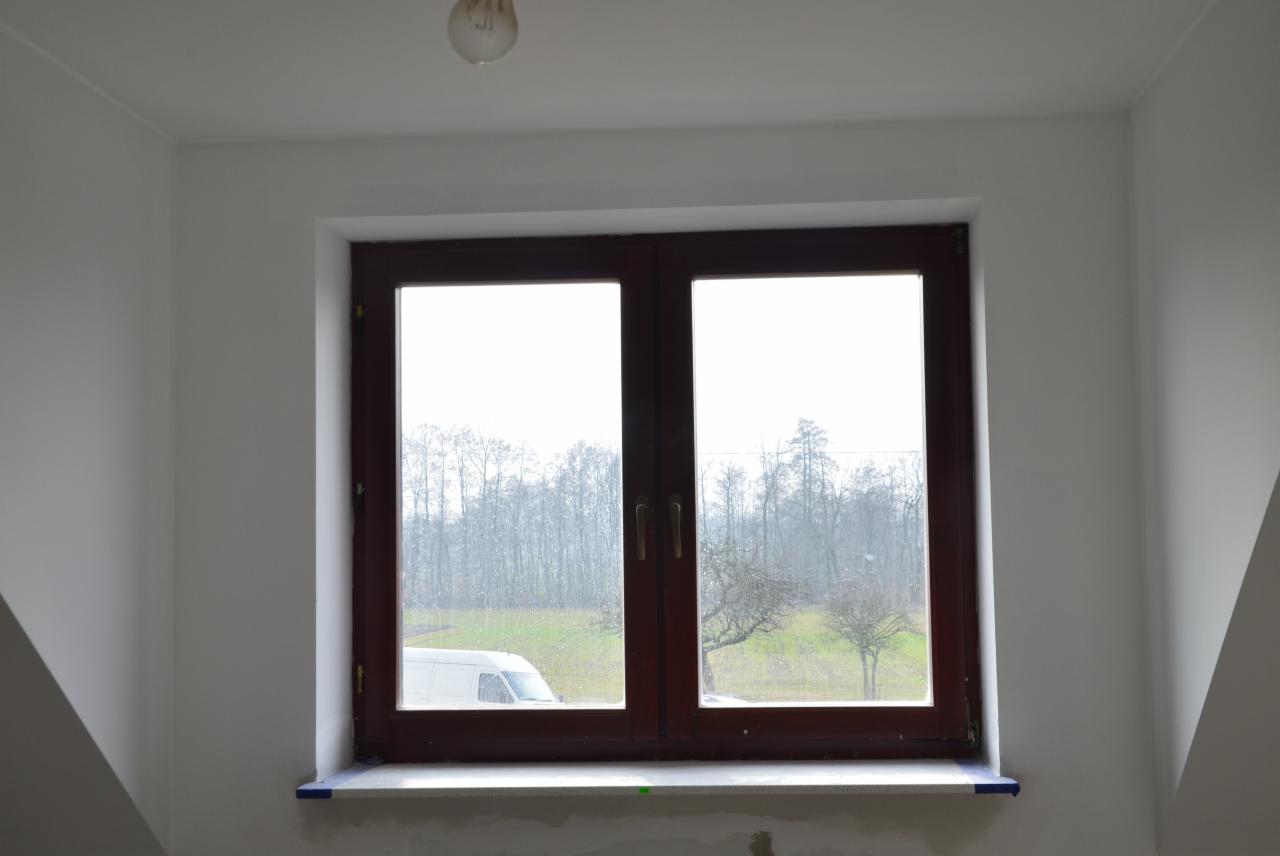 Wnętrze pokoju z oknem, widok na zewnątrz z drzewami i pojazdem dostawczym w tle. Świeżo pomalowane ściany, surowy parapet.