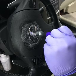 TSW malowanie mycie konstrukcje drewniane remonty - Czyszczenie logotypu na czarnej kierownicy samochodu za pomocą pędzelka, dłoń w fioletowej rękawiczce, widoczny środek czyszczący w butelce.