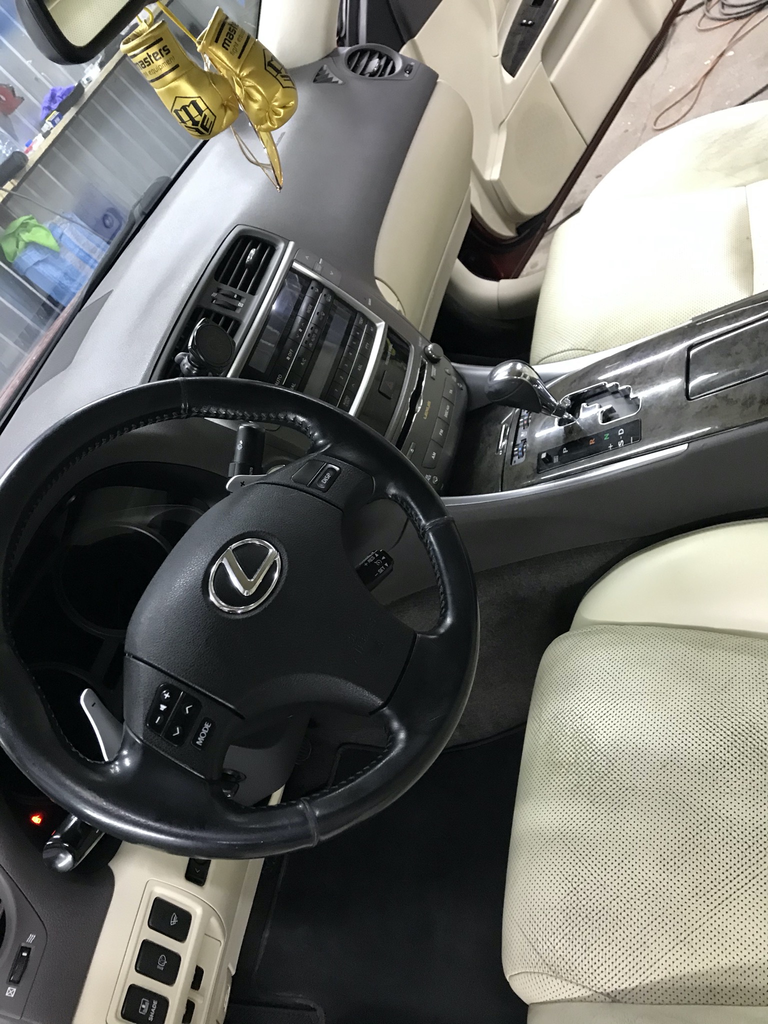 Wnętrze samochodu marki Lexus z jasną skórzaną tapicerką, widok na kierownicę z logo, deskę rozdzielczą i fotele, z ozdobnymi złotymi rękawicami bokserskimi zwisającymi z lusterka.