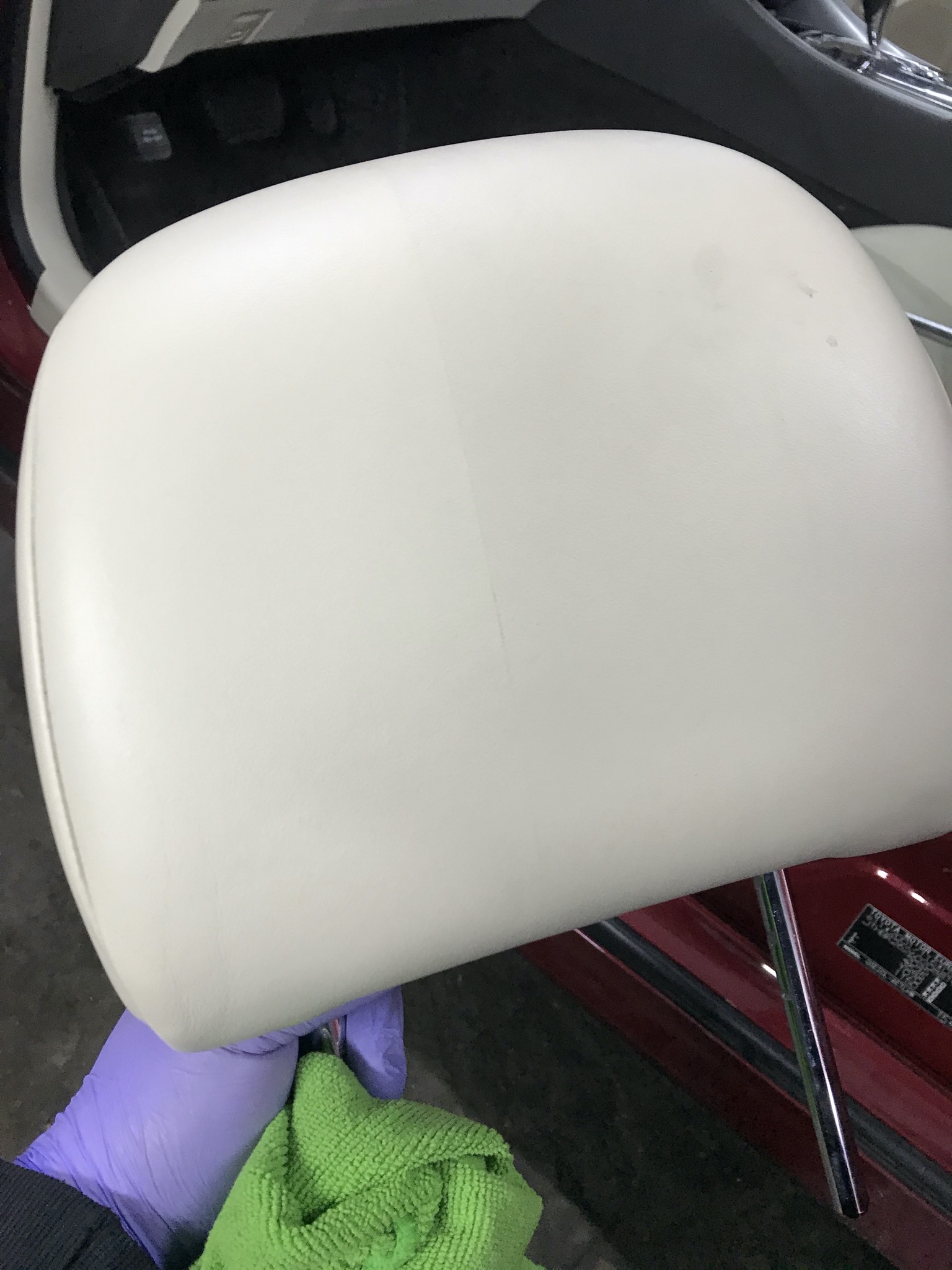 Biały zagłówek samochodowy czyszczony zieloną ściereczką przez osobę w fioletowych rękawiczkach, widoczne wnętrze samochodu.