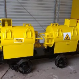 Żółty wagonik do transportu substancji łatwopalnych z tabliczką ostrzegawczą, na czarnych kołach, część większego zestawu w hali z blachy falistej.