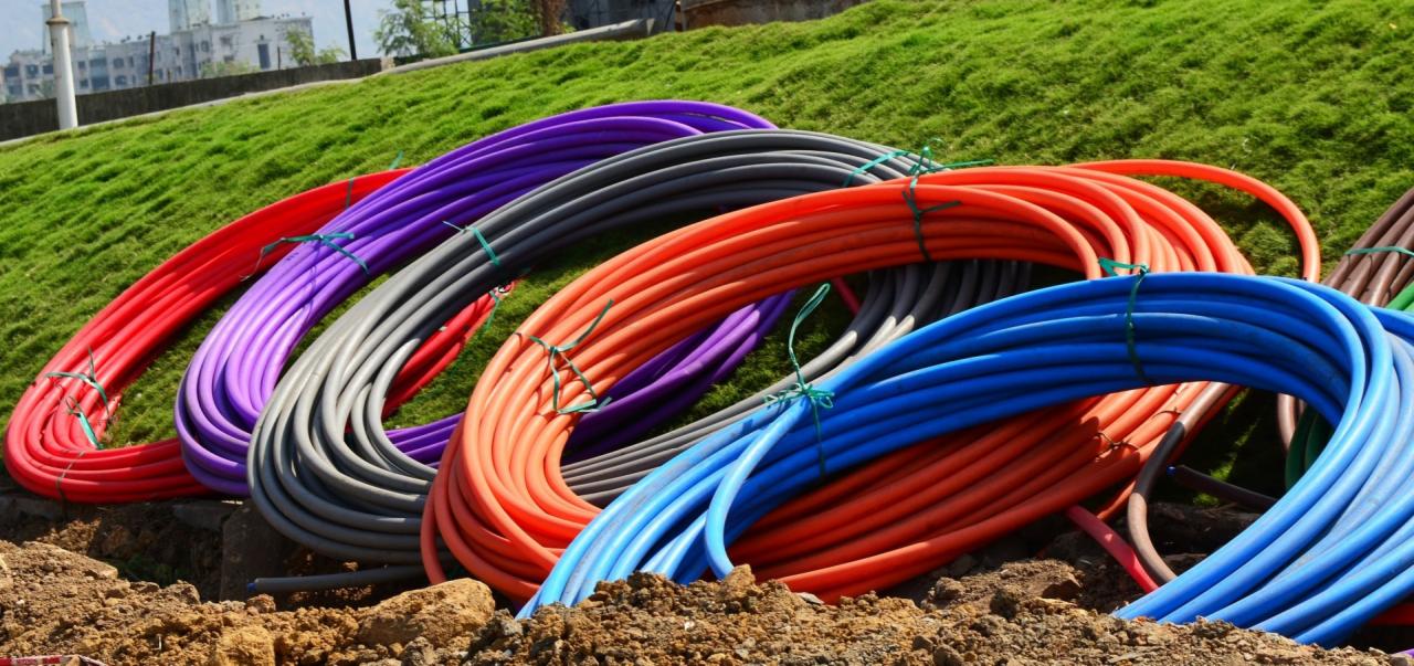 Kolorowe zwoje rur (czerwona, fioletowa, szara, pomarańczowa, niebieska) leżą na ziemi z zieloną trawą w tle. Rury związane zielonymi sznurkami.