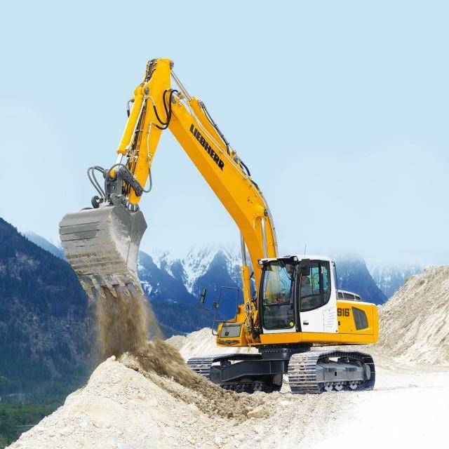 Żółta koparka Liebherr 916 nabiera ziemię na tle gór. Maszyna na gąsienicach wykonuje prace ziemne, widoczne świeże ślady. Jasne, naturalne oświetlenie.