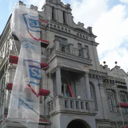 Renowacja elewacji zabytkowego budynku z rusztowaniami i siatką ochronną. Widoczne detale architektoniczne i fragment błękitnego nieba.