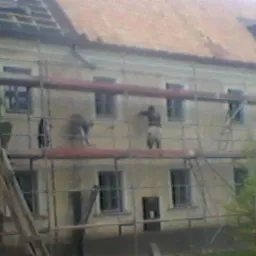 Praca na rusztowaniu przy elewacji dwupiętrowego budynku z czerwonym dachem. Widoczne okna i fragment dachu z lukarną. Częściowo odsłonięta konstrukcja dachu.