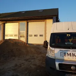 Biała brama garażowa segmentowa z czterema małymi okienkami, widoczna z perspektywy ziemi, obok zaparkowany biały bus z logo firmy EMKAT Salon, na pierwszym planie pryzma piasku.