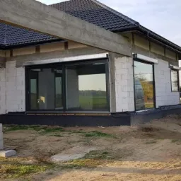 Dom w trakcie budowy z zamontowanymi oknami z czarnymi ramami i roletami zewnętrznymi, obok zaparkowany biały bus dostawczy.