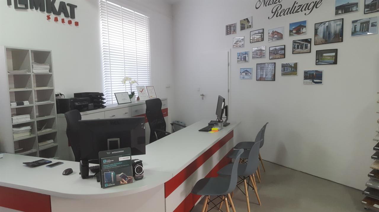 Wnętrze biura EMKAT Radomsko z ekspozycją rolet wewnętrznych za biurkiem i galerią zdjęć z realizacji na ścianie.