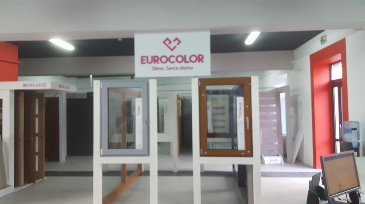 Ekspozycja modeli okien w salonie, widoczne okna w kolorach szarym i brązowym na białych stojakach, w tle logo firmy Eurocolor i inne ekspozycje.