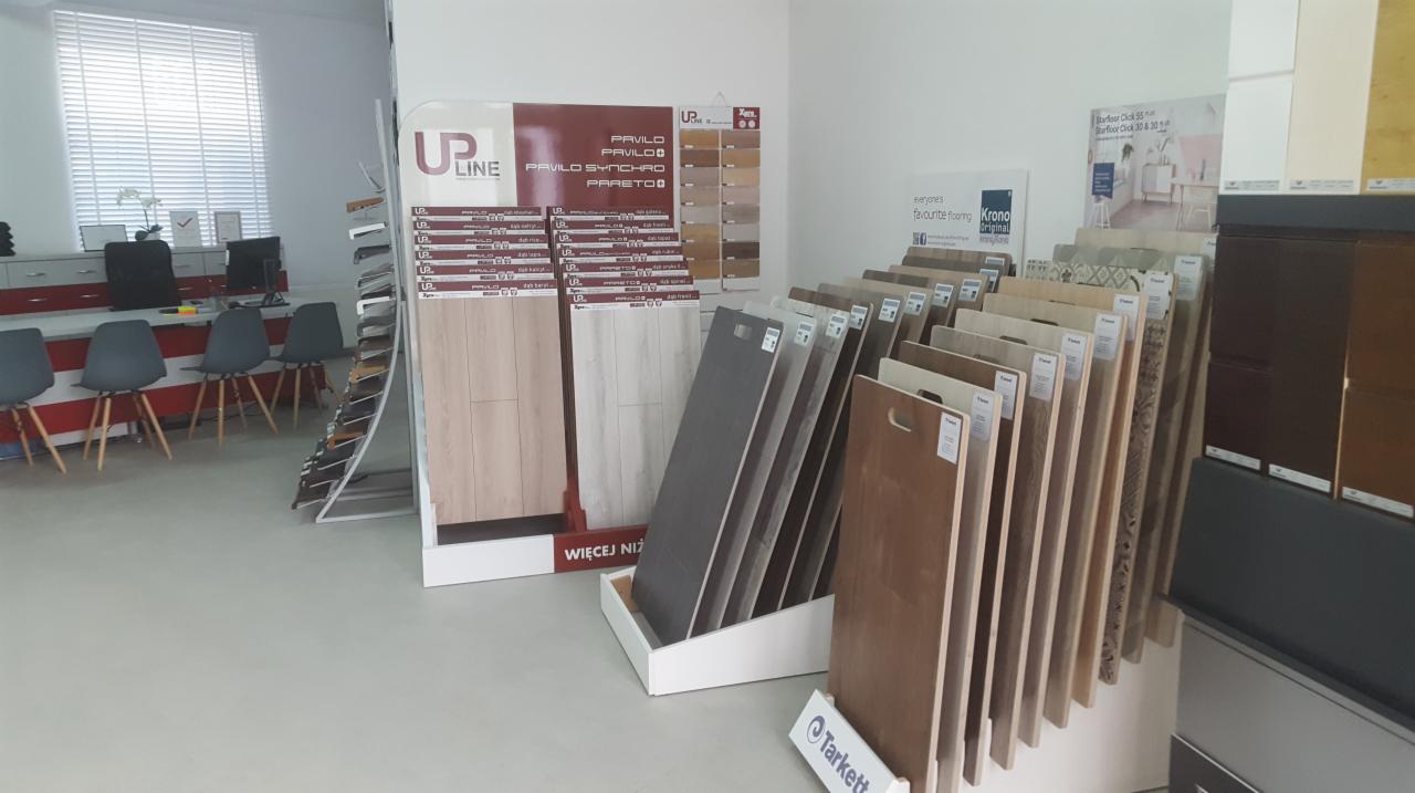 Ekspozycja próbek paneli podłogowych różnych producentów i wzorów w salonie sprzedaży, widoczne stojaki z panelami, logo Tarkett na jednym ze stojaków, w tle biurko i krzesła dla klientów.