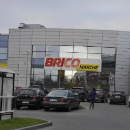 Fasada sklepu Brico Marche z dużym logo, parkingiem i kilkoma samochodami. Widoczna osoba przechodząca przez parking. Budynek o nowoczesnej architekturze.