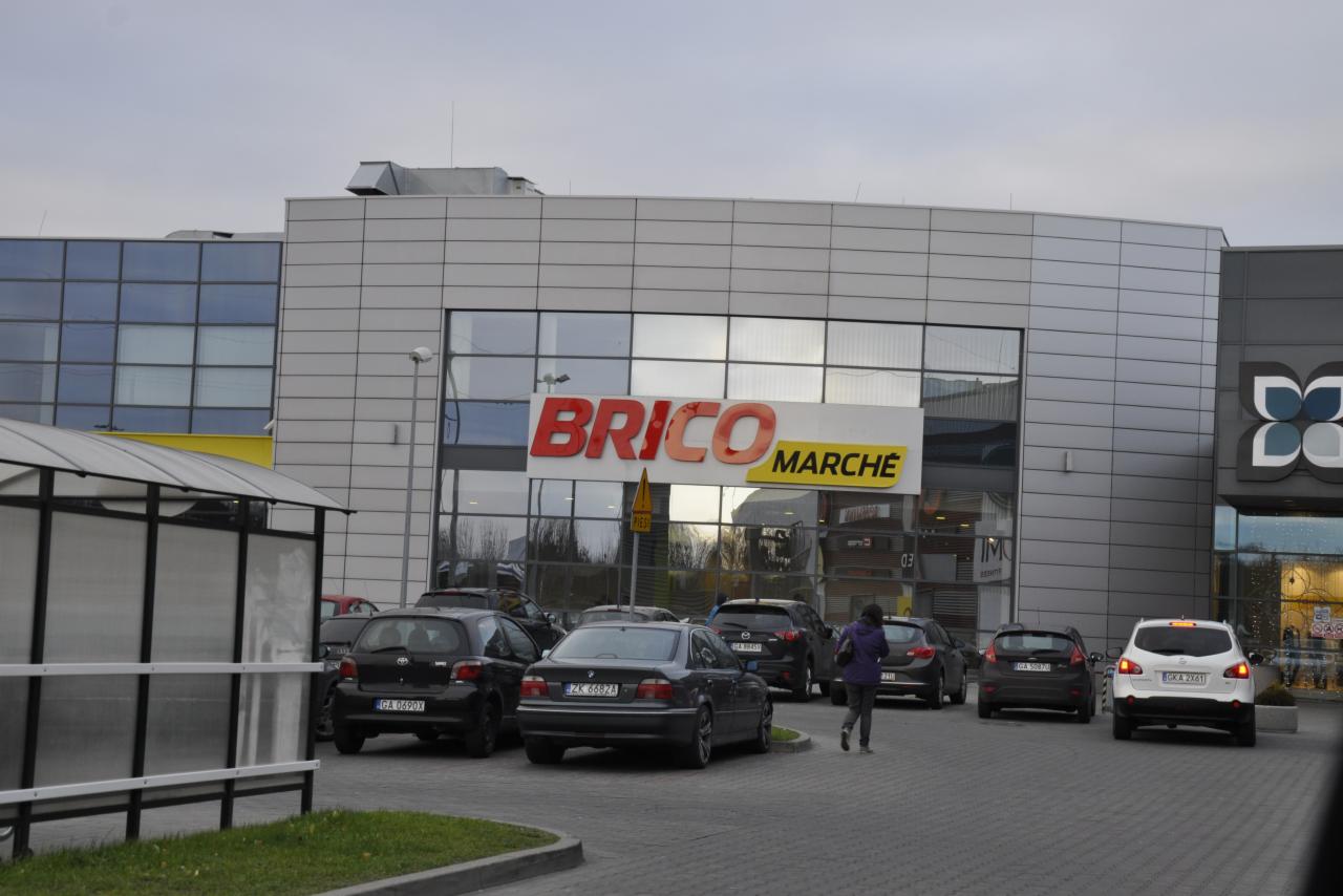 Fasada sklepu Brico Marche z dużym logo, parkingiem i kilkoma samochodami. Widoczna osoba przechodząca przez parking. Budynek o nowoczesnej architekturze.