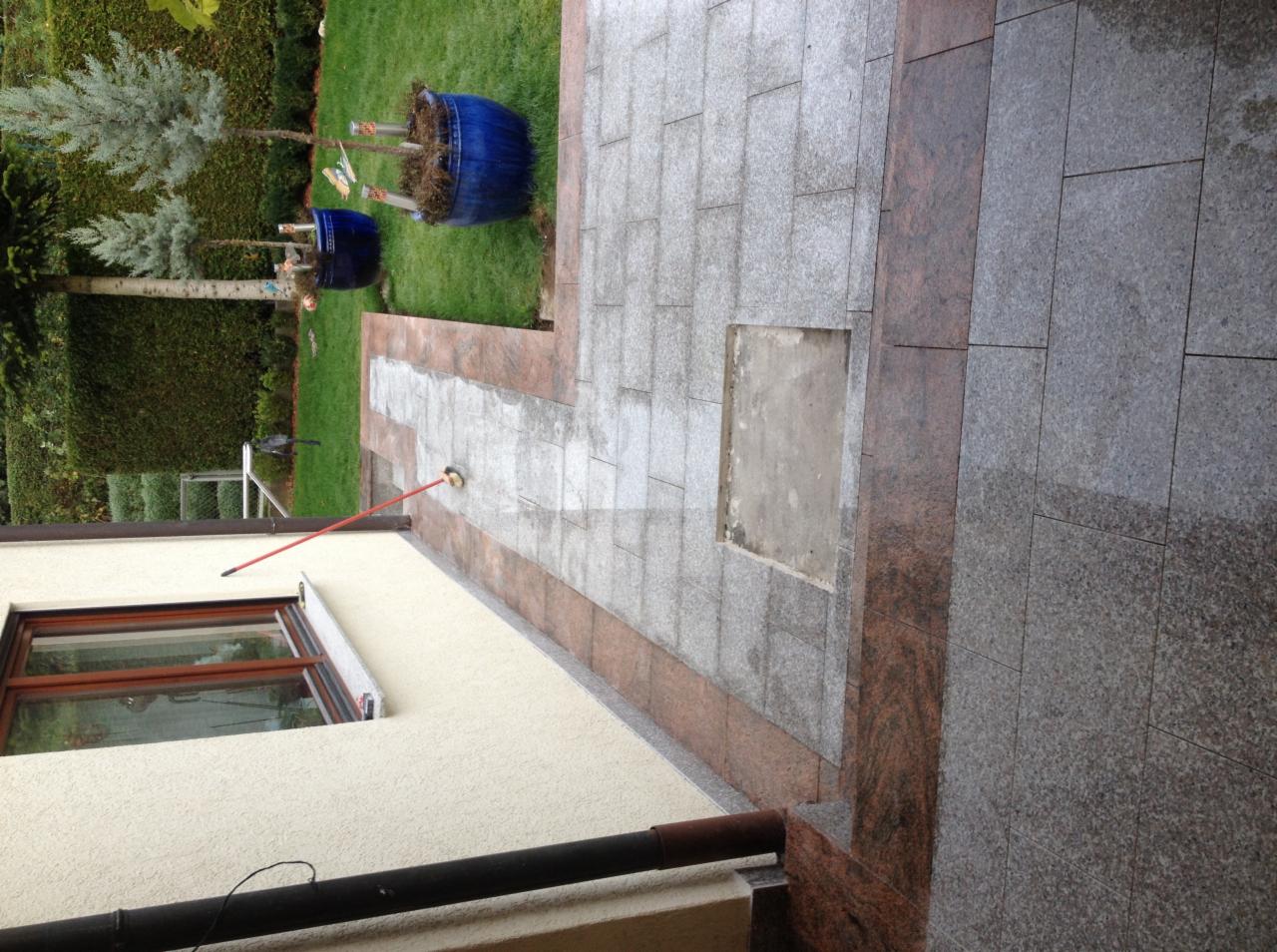 Ułożona szara kostka brukowa z bordową obwódką na tarasie, widoczne niebieskie donice z roślinami i fragment elewacji budynku z oknem. Kostka jest mokra, prawdopodobnie po myciu.