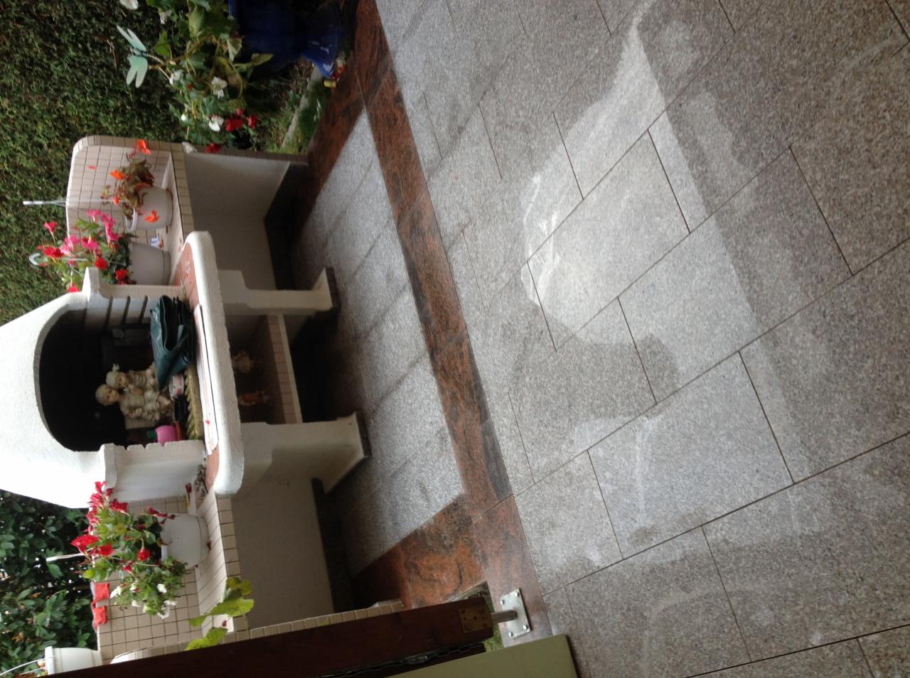 Ułożona, szara, prostokątna kostka brukowa z bordową obwódką. Z lewej strony grilla z kwiatami. Całość sfotografowana pod kątem.