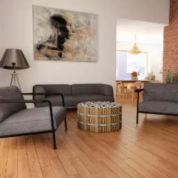 Nowoczesne wnętrze salonu z szarą sofą, fotelami z czarną ramą, okrągłym stolikiem z drewnianych bali i abstrakcyjnym obrazem na ścianie, oświetlone lampą stojącą z metalowym abażurem.