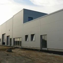 Nowoczesna hala magazynowa z blachy trapezowej w trakcie budowy, widok z zewnątrz. Szara elewacja, okna i brama garażowa. Teren przygotowany pod wylewkę.