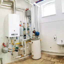 Biały kocioł gazowy Prestige z podłączonymi rurami miedzianymi i zbiornikiem wyrównawczym w pomieszczeniu z białą umywalką i zmiękczaczem wody.