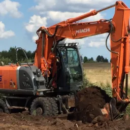 Pomarańczowy kołowy koparka Hitachi w trakcie prac ziemnych na polu, z widocznym nabranym urobkiem w łyżce. Błękitne niebo z chmurami w tle.