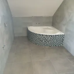 Wykończona łazienka na poddaszu z narożną wanną obłożoną mozaiką czarno-białą, szare płytki na ścianach i podłodze, widoczne niezamaskowane przyłącza wodne.