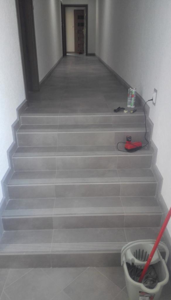 Wykończone schody i podłoga w korytarzu pokryte szarymi płytkami. Widoczne narzędzia glazurnicze i wiadro z mopem. Płytki schodowe z antypoślizgowymi paskami.