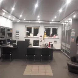 Wnętrze biura z ekspozycją okien i drzwi. Widoczne biurka, krzesła, okna, drzwi przesuwne, sofa i makieta mężczyzny. Oświetlenie sufitowe.