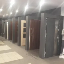 Ekspozycja różnorodnych drzwi zewnętrznych w salonie. Widoczne modele w różnych kolorach i stylach, ustawione w rzędzie. W tle regały z produktami.