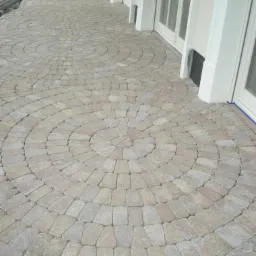 Ułożona z beżowo-różowych kostek nawierzchnia w kształcie koncentrycznych kręgów, przylegająca do białej elewacji budynku z oknami.