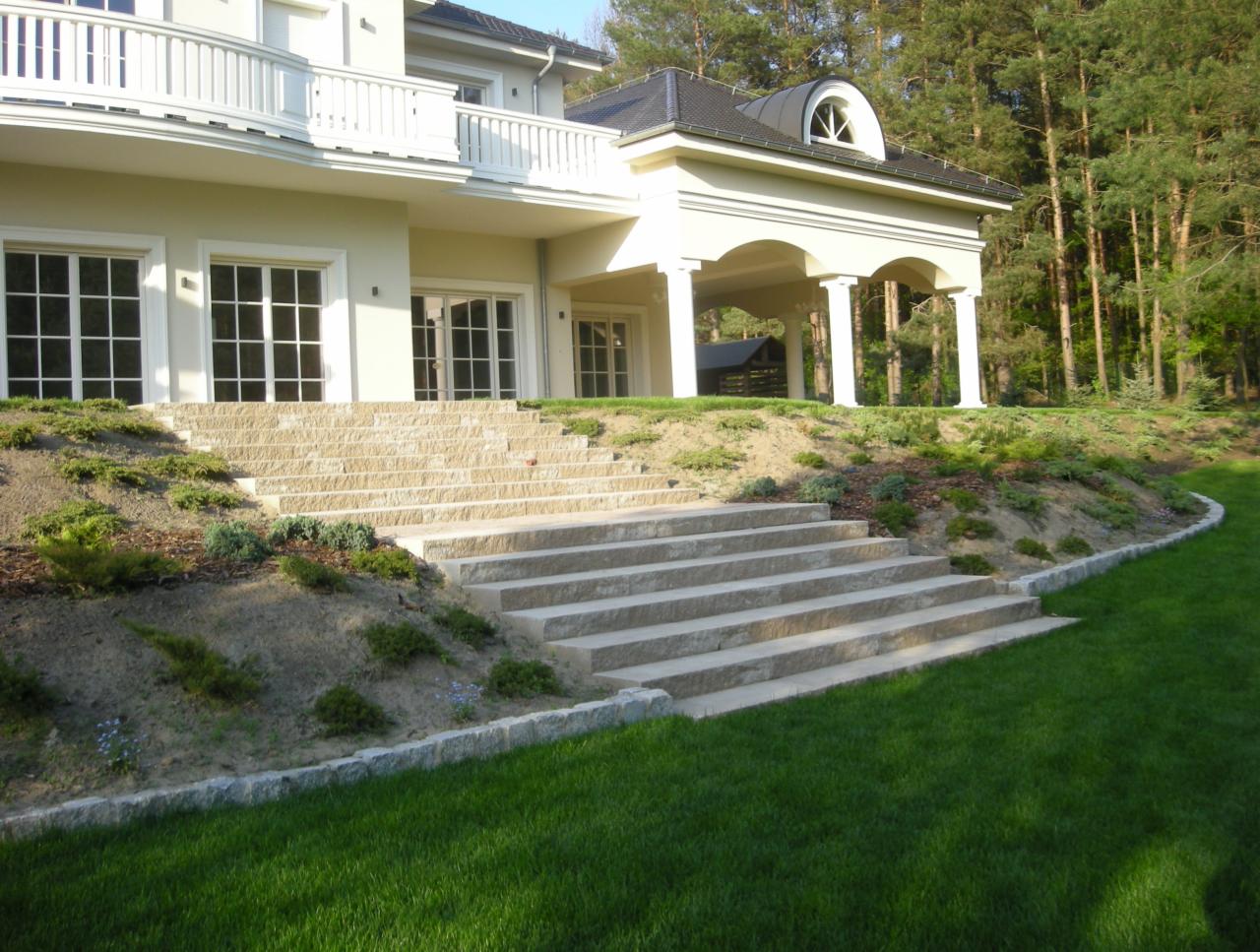 Kamienne schody prowadzą do domu z ogrodem, otoczone zielenią i niskimi krzewami. Widoczny trawnik i drzewa w tle. Schody wykonane z jasnego kamienia.
