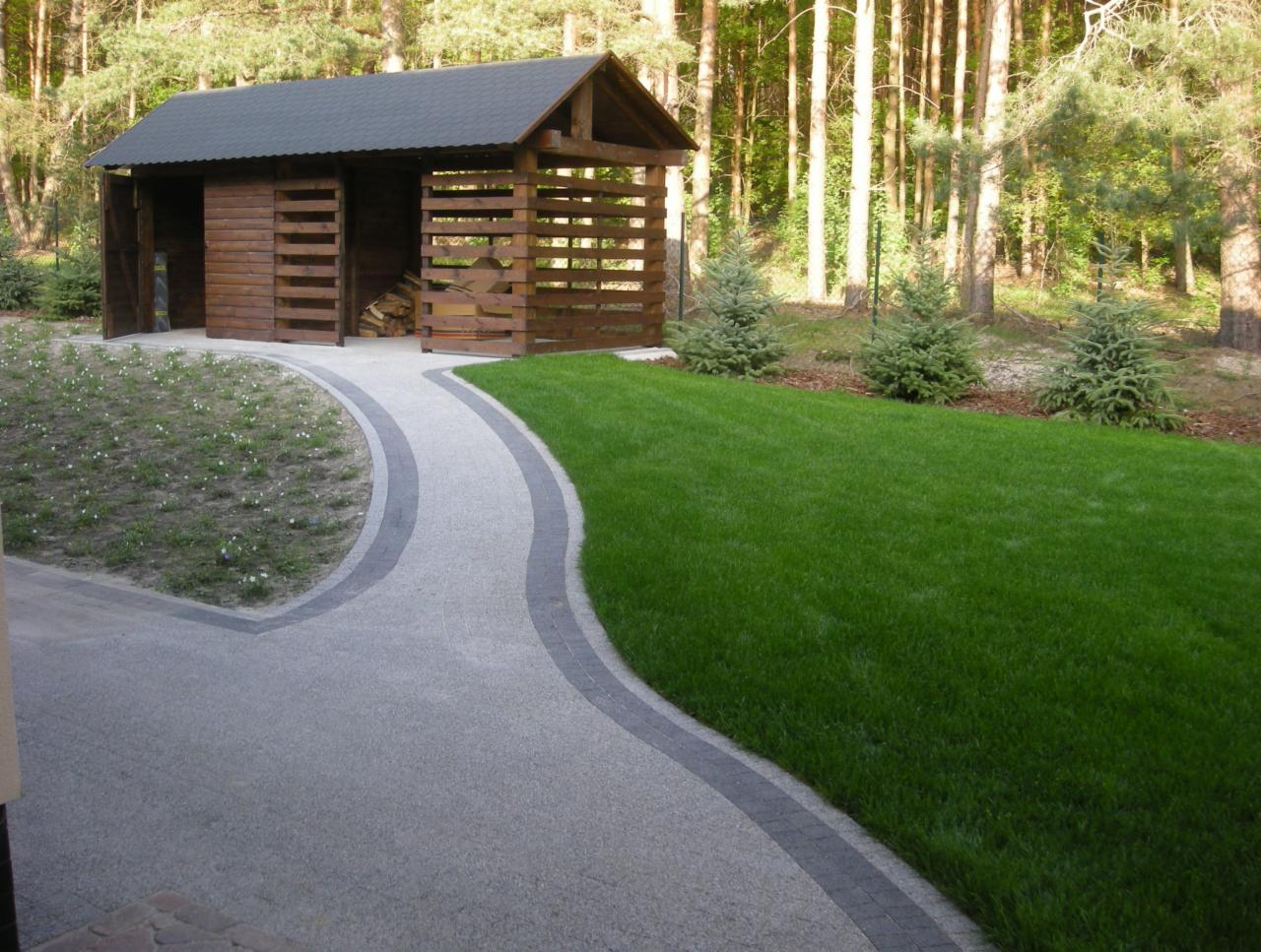 Ścieżka z jasnej kostki prowadzi do drewnianej drewutni na tle lasu. Po prawej stronie widoczny fragment trawnika i małe drzewka iglaste.