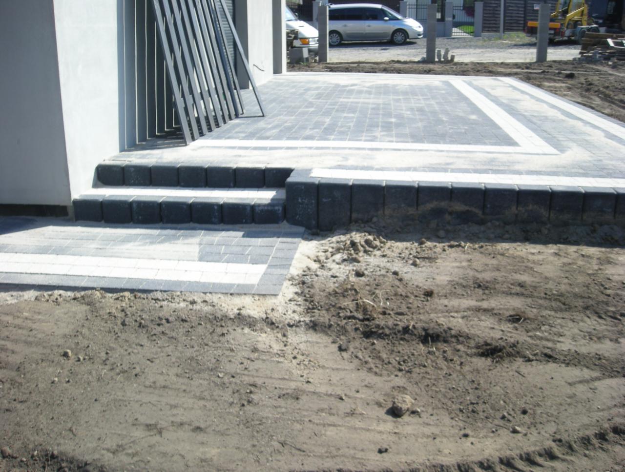 Ułożona kostka brukowa w szaro-białej kolorystyce tworzy wzór na podjeździe. Widoczne schody z kostki i niezabudowany teren wokół.