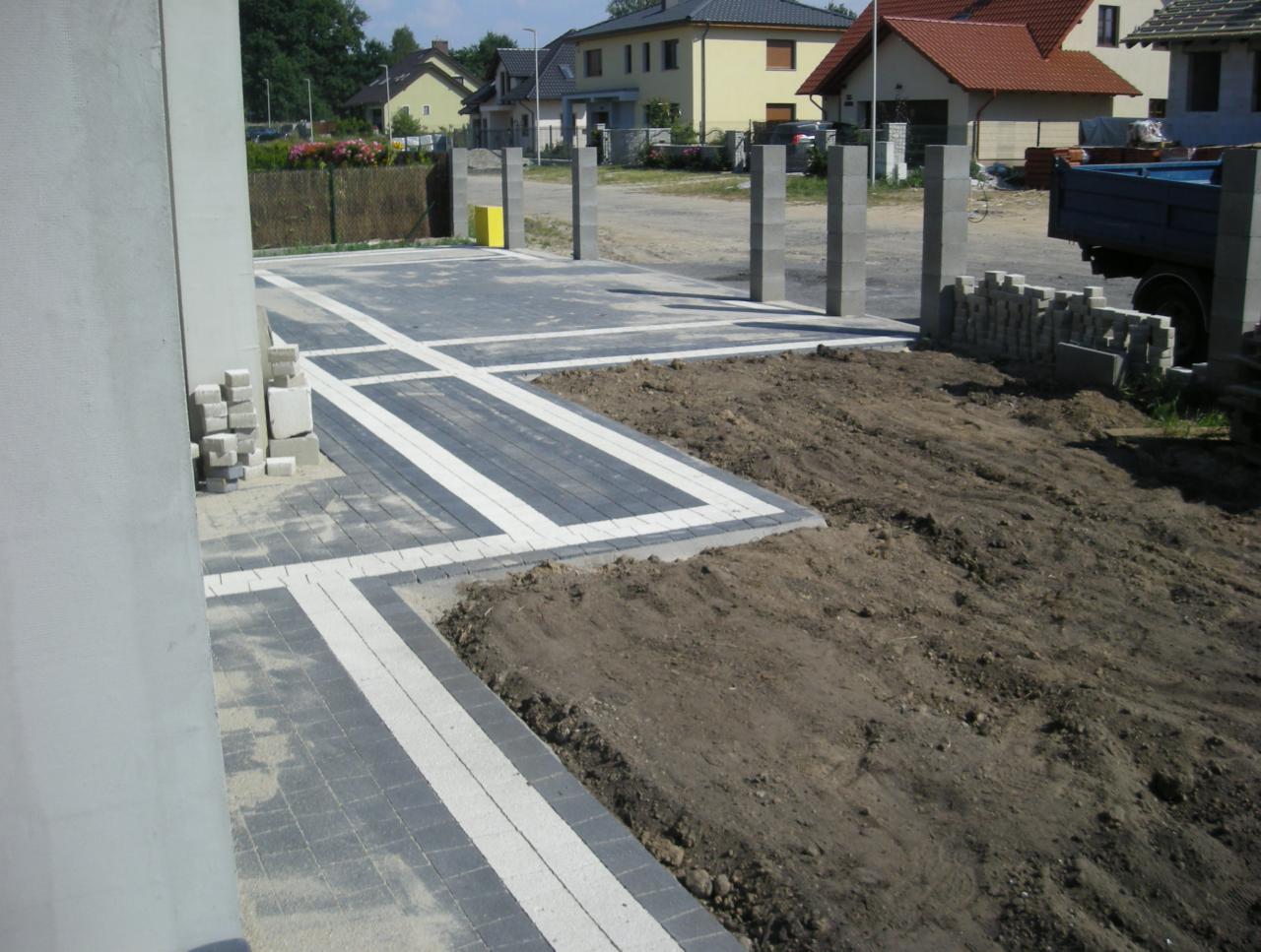 Częściowo ułożona, dwukolorowa kostka brukowa z białymi akcentami, na podjeździe. W tle widoczne słupki ogrodzeniowe i przygotowany teren pod dalsze prace.