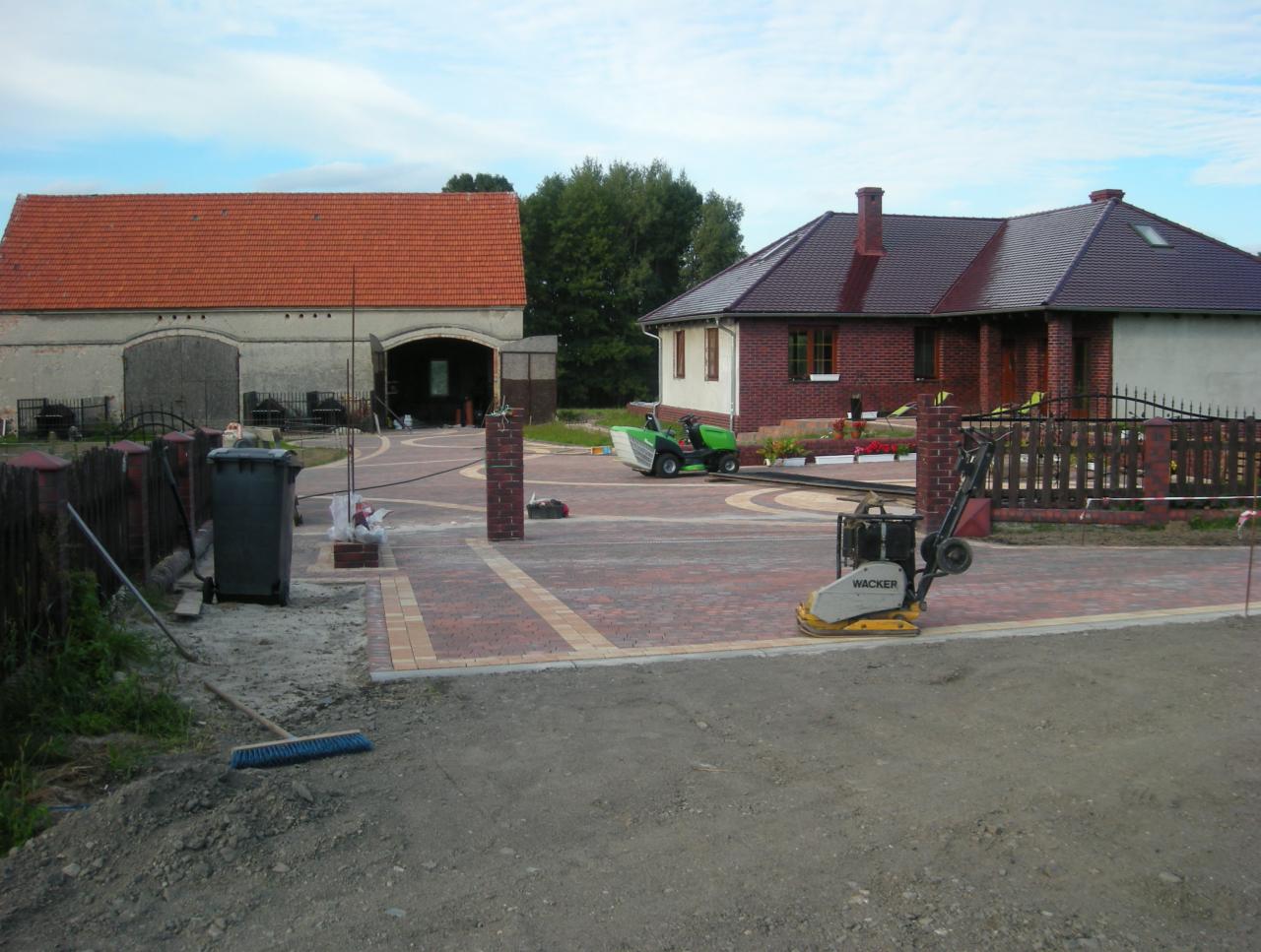 Rozległy podjazd z czerwono-beżowej kostki, oddzielony ceglanymi słupkami. W tle budynek gospodarczy i dom z czerwonej cegły. Widoczna zagęszczarka Wacker.