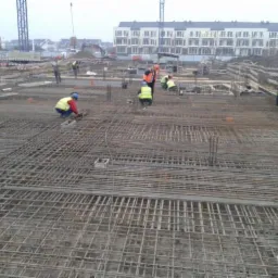 Zbrojenie fundamentów na budowie z pracownikami w odblaskowych kamizelkach. W tle budynki w stanie surowym i żurawie budowlane. Praca wre na dużej powierzchni.