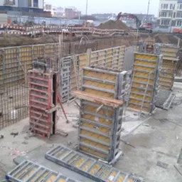 Konstrukcja szalunkowa na placu budowy, widoczne zbrojenia i przygotowania do wylewania fundamentów. W tle budynki mieszkalne i koparka.