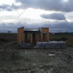 Wznoszenie ścian z czerwonej cegły na budowie, widoczne palety z materiałami i taczka, pochmurne niebo w tle, teren budowy w trakcie realizacji.