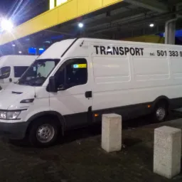 Biały samochód dostawczy z napisem 'TRANSPORT' i numerem telefonu zaparkowany na zewnątrz, widoczne dwa betonowe słupki i drugi samochód w tle, oświetlenie uliczne.