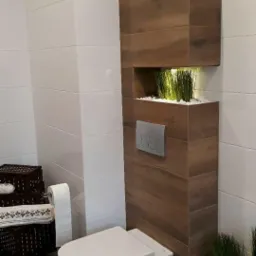 Nowoczesna łazienka z wiszącą toaletą, drewnianą kolumną z podświetlonymi niszami wypełnionymi zielenią i białymi kamykami, białe płytki na ścianach i brązowe na podłodze.