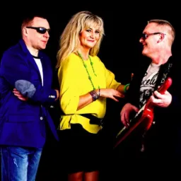 Trzyosobowy zespół: mężczyzna w niebieskiej marynarce i jeansach, kobieta w żółtej bluzce i czarnej spódnicy oraz mężczyzna z czerwoną gitarą Roland, wszyscy uśmiechnięci na czarnym tle.
