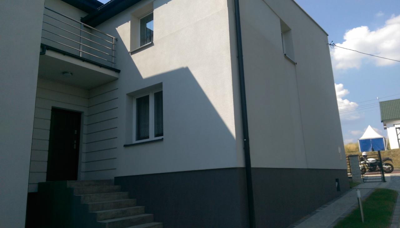 Otynkowany budynek jednorodzinny z balkonem i widocznymi schodami wejściowymi. Elewacja w odcieniach szarości. W tle błękitne niebo i częściowo widoczny motocykl.